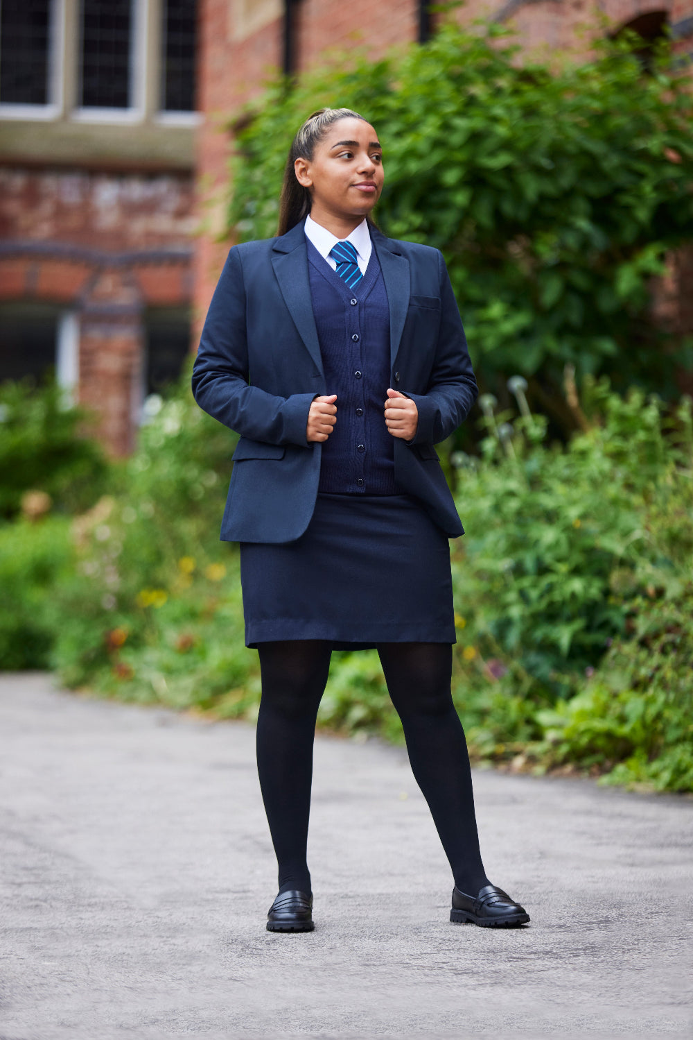 Schoolgirl wearing a navy blue uniform standing outdoors with greenery and a brick building in the background